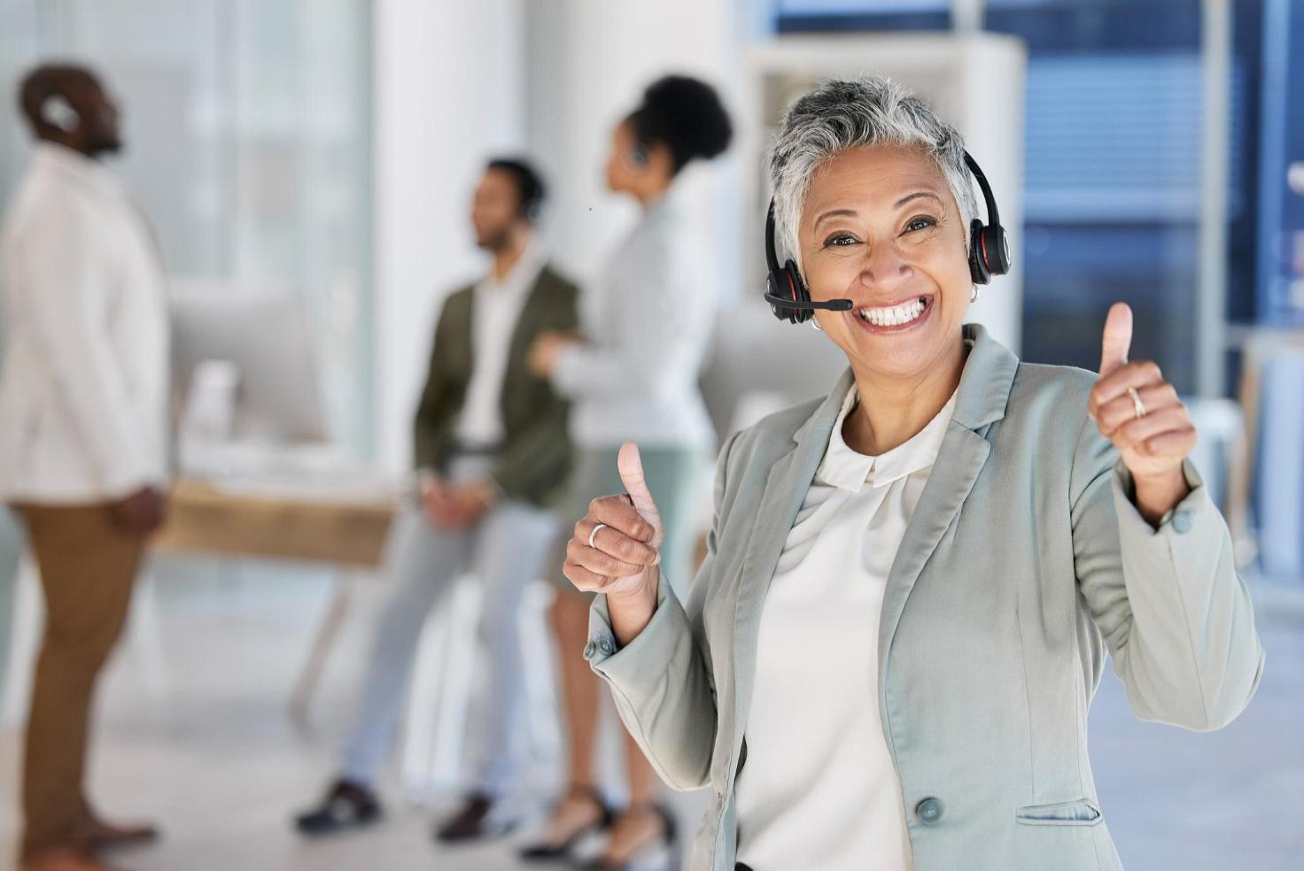Woman support agent thumbs upping in cloud contact center with team in background, all wearing headsets.